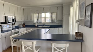 A kitchen with white cabinets, a blue countertop, a central island with two white chairs, a microwave, a stove, and a sink. - 
Cozy Waterfront Beach House with Private Beach/Pier/Tennis Courts (Pensacola)