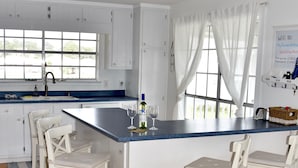 A modern kitchen with white cabinetry, a central island with bar stools, a sink, and a window with curtains. - 
Cozy Waterfront Beach House with Private Beach/Pier/Tennis Courts (Pensacola)