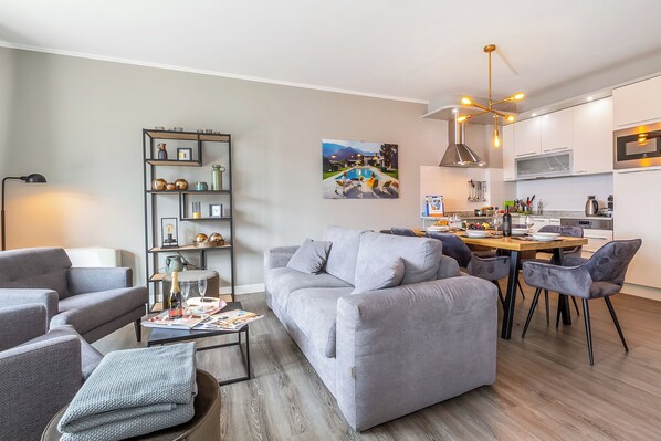 A modern living room with a grey sofa, a coffee table with magazines, and a dining area with a wooden table and chairs. - Porto Letizia P8, Porlezza, Italy (Porlezza)