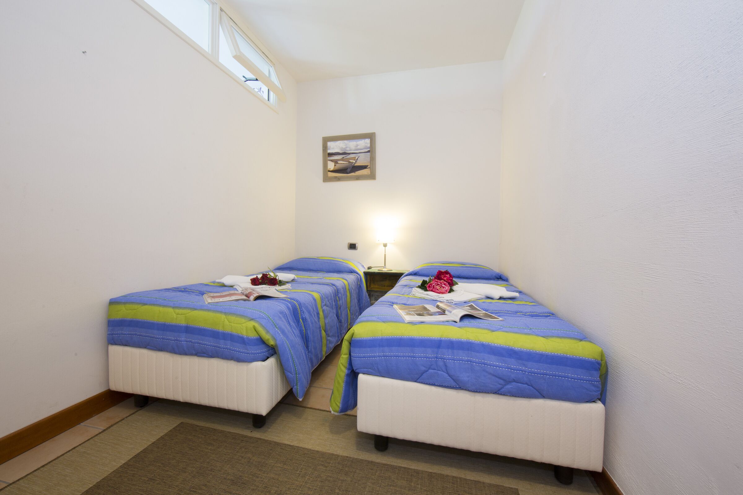 A small room with two single beds, each with a blue and green striped bedspread, a framed picture on the wall, and a nightstand with a lamp.