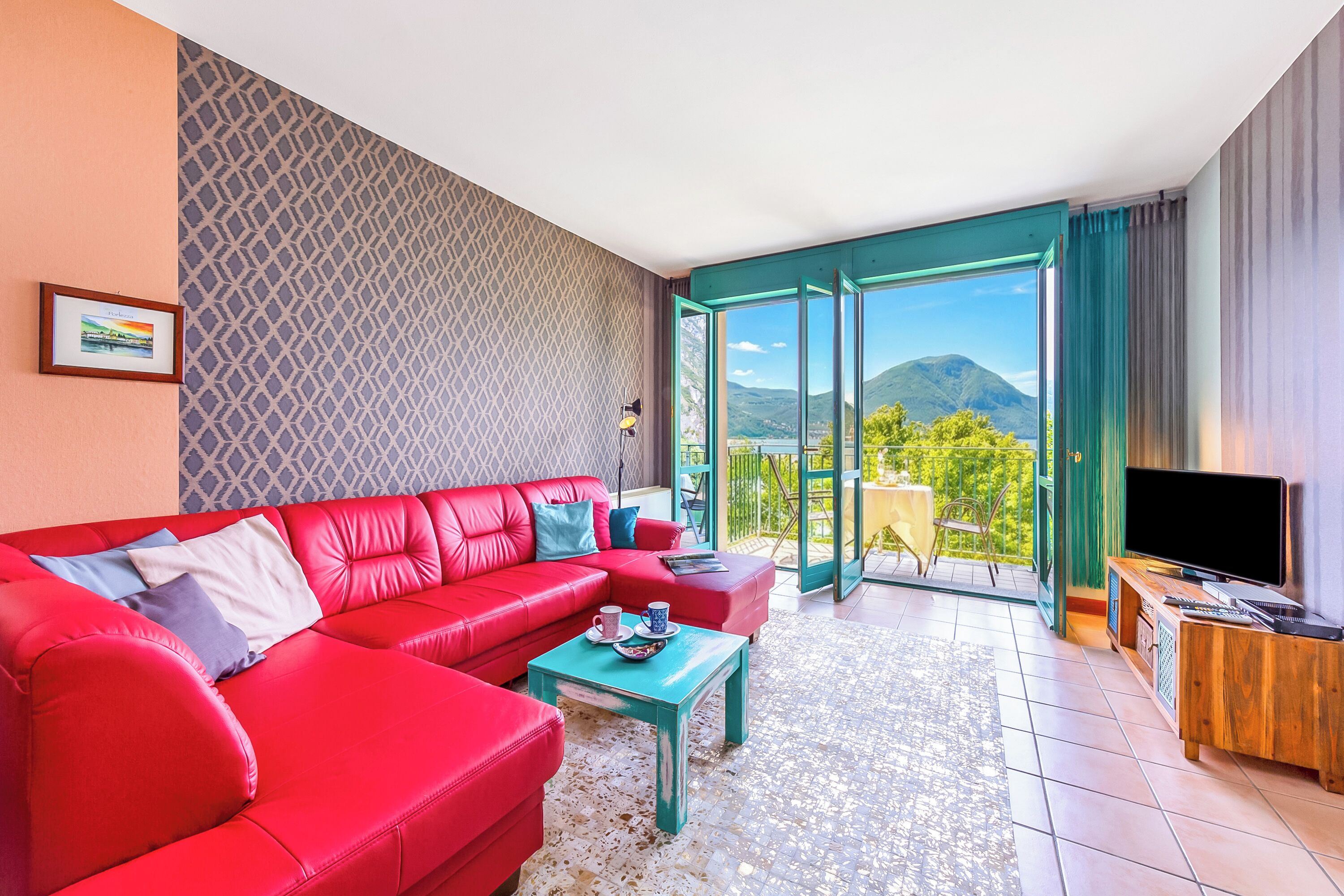 A red sofa, a teal coffee table, a TV, and a balcony with a mountain view.