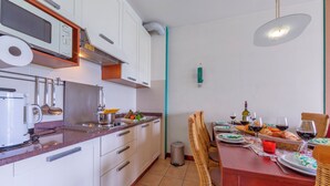 Pieni, käytännöllinen keittiö, jossa on valkoiset kaapit, mikroaaltouuni ja pesuallas. Ruokapöytä on katettu ja sen ympärillä on tuoleja, ja pöydällä on viinilaseja ja -pulloja.