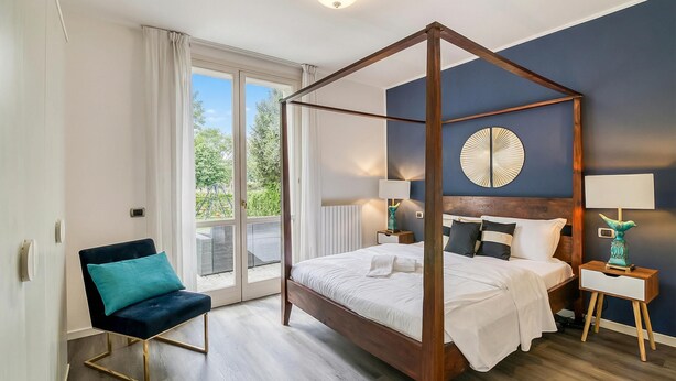 A bedroom with a four-poster bed, a chair, bedside tables, and a view through a large window. - Porto Letizia Orchidea 2, Porlezza, Italy (Porlezza)
