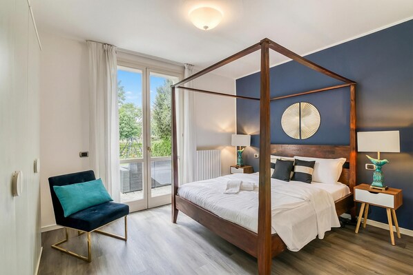 A bedroom with a four-poster bed, a chair, bedside tables, and a view through a large window.