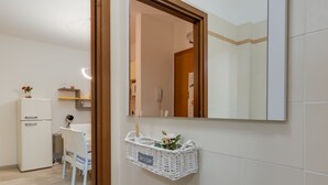 Et badeværelse med en hvid flisevæg, et spejl med træramme, en fletkurv med etiket, en hvid stol og en hvid radiator.