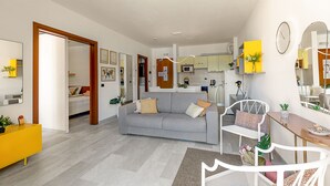 En moderne stue med en grå sofa, et gult sofabord og en hvid stol. Et køkken kan ses i baggrunden med en mikrobølgeovn og skabe.