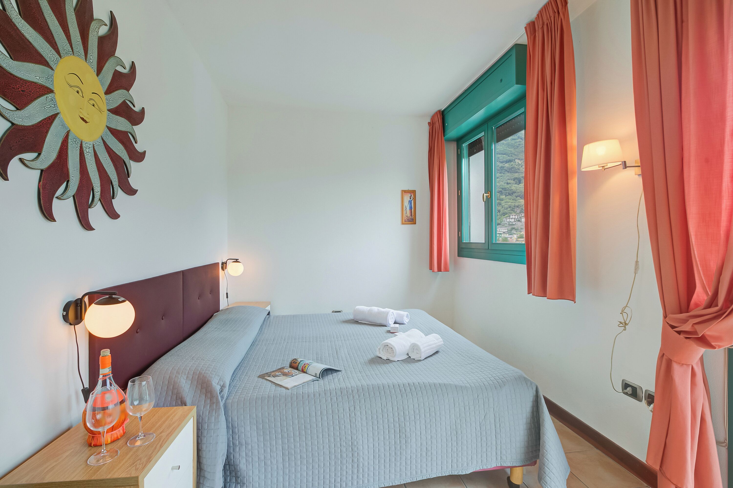 A bedroom with a bed, a nightstand with a lamp, a window with curtains, and a wall decoration.