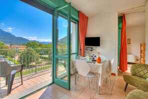 Uma varanda com mesa posta para refeições, uma televisão fixada na parede e vista para montanhas e casas.
