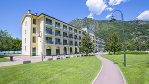 A multi-story hotel with a central courtyard, a paved walkway, and a mountain backdrop. - Porto Letizia Ortensia 3, Porlezza, Italy (Porlezza)