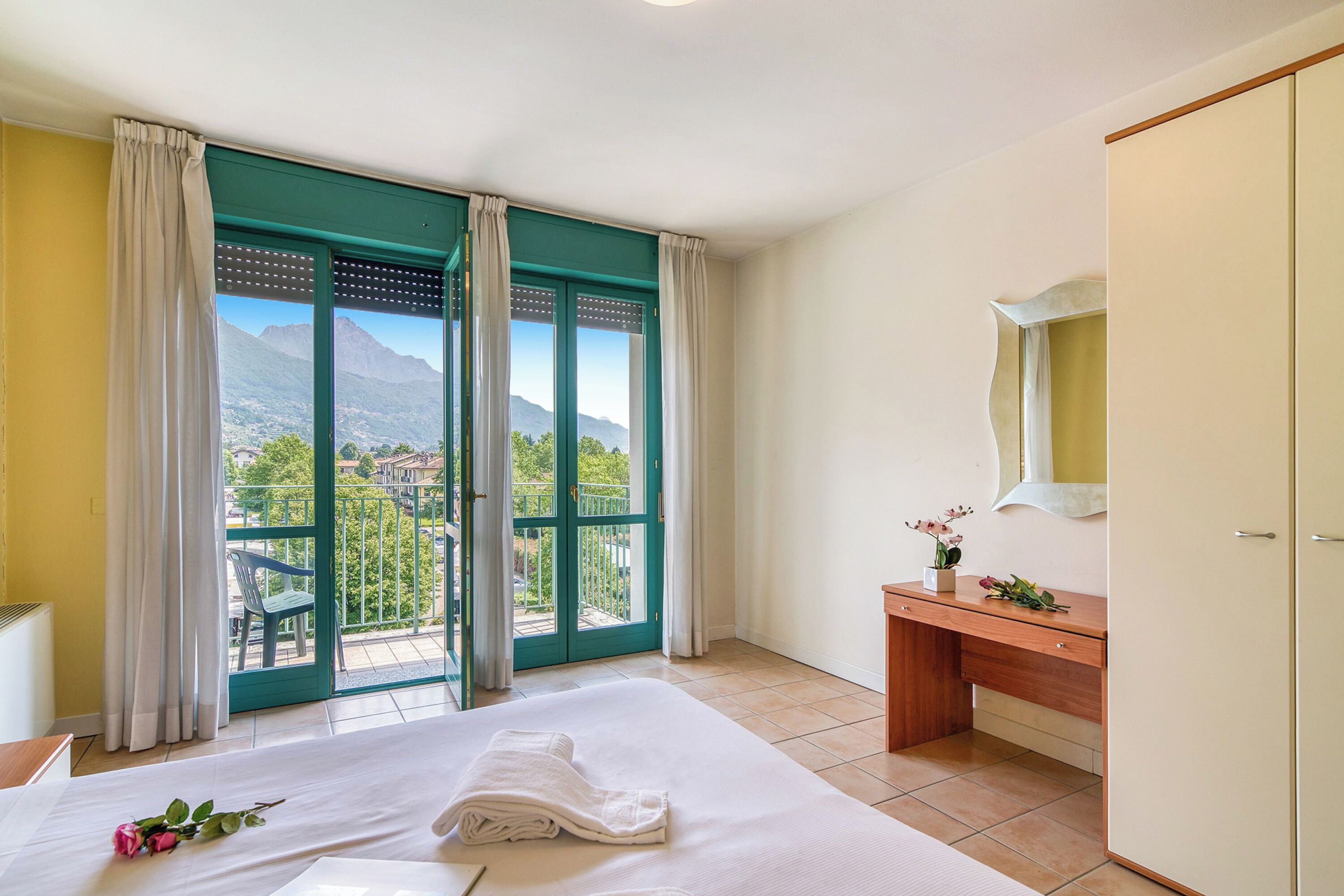 Une chambre d’hôtel équipée d’un lit, d’un bureau, d’un miroir et d’un balcon offrant une vue sur les montagnes.