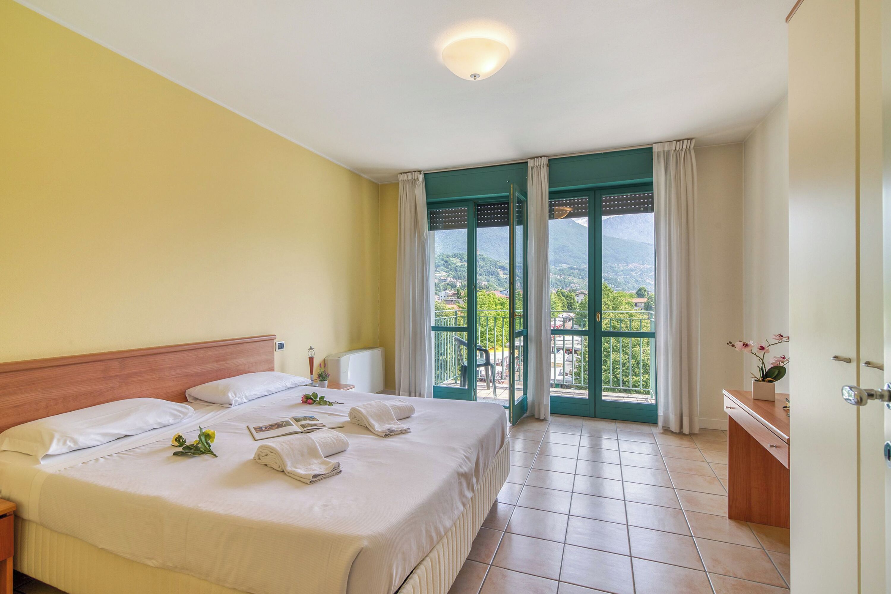 A hotel room with a large bed, a wooden headboard, a bedside table with a lamp, and a view of greenery through the open door.