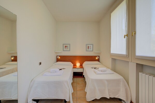 Quarto com duas camas de solteiro, mesa de cabeceira com abajur, espelho e janela com persianas.