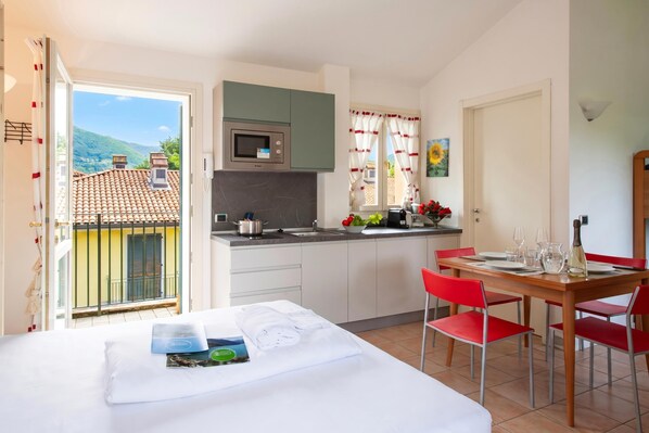 A hotel room with a bed, a dining table with red chairs, a kitchen with a microwave, and a view of the mountains. - Porto Letizia Mimosa 5, Porlezza, Italy (Porlezza)
