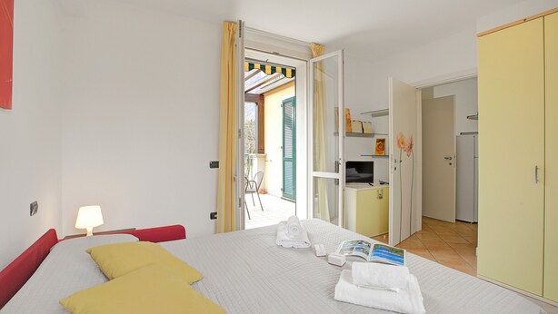 Habitació d'hotel amb un llit, una tauleta de nit amb una làmpada, un armari i una porta oberta cap a un balcó.