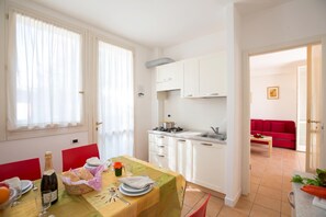 Eine moderne Küche mit weißen Schränken, ein Essbereich mit einem Tisch für Zwei und ein rotes Sofa im Wohnzimmer.