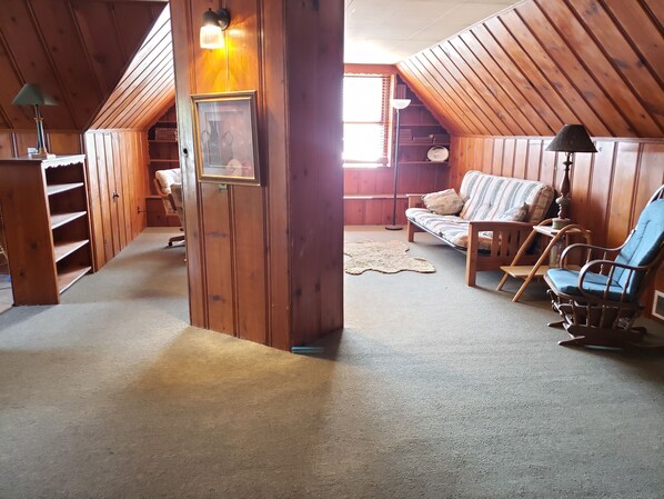 A cozy attic room with wooden walls, a framed picture, a sofa, a rocking chair, and a lamp.
