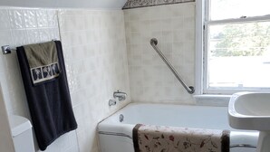 Een badkamer met een wit toilet, een bad met een bloemenhanddoek, een witte wastafel en een raam.
