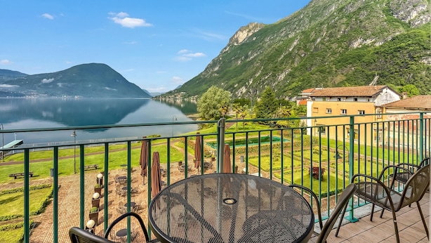 Un balcon avec vue sur un lac et des montagnes, des sièges d’extérieur et un bâtiment en arrière-plan.