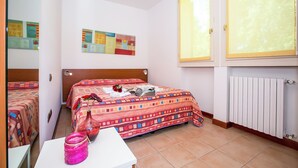 A bedroom with two beds, a white table, a pink vase, and a window with blinds. - Porto Letizia Dalia 4, Porlezza, Italy (Porlezza)