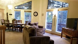 A living room with a brown sofa, a dining table with chairs, a TV stand, and large windows with a view of trees. - Eagle Cliff Cottage  Sleeps 6 ((20-NCD0244) (Estes Park)