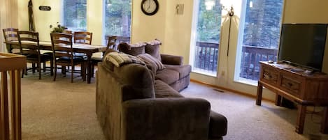 Una sala de estar con un sofá marrón, una mesa de comedor con sillas, un mueble para la televisión y ventanas amplias con vista a árboles.