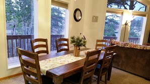 A dining area with a wooden table and chairs, a clock on the wall, and large windows with a view of trees. - Eagle Cliff Cottage  Sleeps 6 ((20-NCD0244) (Estes Park)