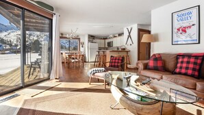 Un vaste espace de vie comprenant une table basse en verre, un canapé en cuir avec des coussins rouges et noirs, et un coin repas avec des chaises en bois. On aperçoit une cuisine au fond, et une grande fenêtre offre une vue sur un paysage enneigé.