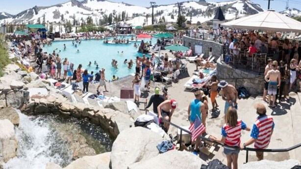 Une piscine extérieure très fréquentée, où les gens se baignent et profitent de l’eau, le tout dans un cadre magnifique, entouré de montagnes et d’un paysage rocheux.