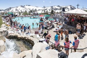Une piscine extérieure très fréquentée, où les gens se baignent et profitent de l’eau, le tout dans un cadre magnifique, entouré de montagnes et d’un paysage rocheux.
