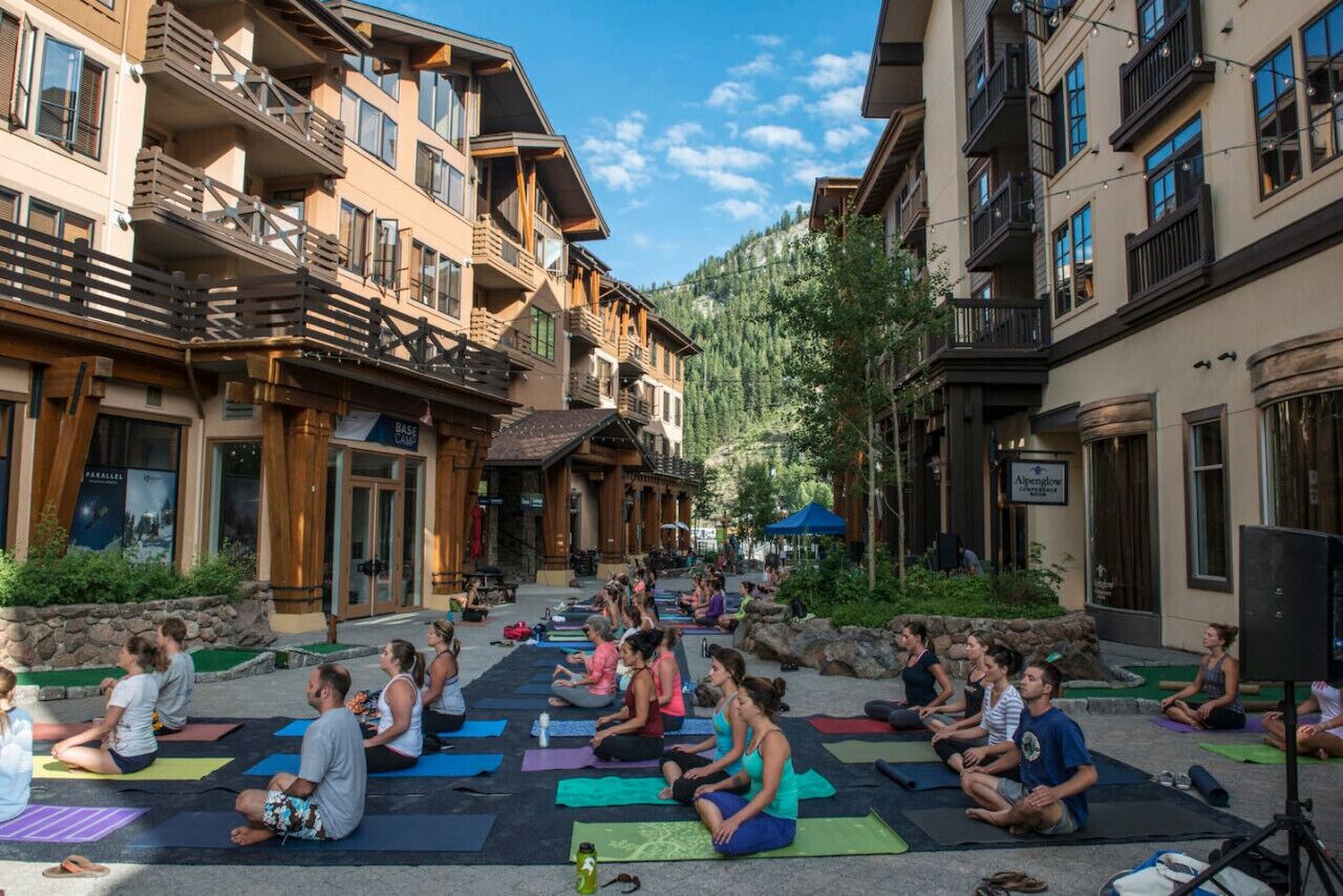 A group of people sitting on yoga mats in an outdoor setting with buildings in the background.