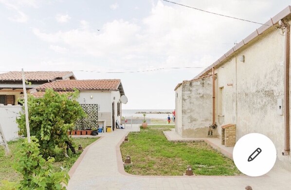 Un étroit sentier mène à un bâtiment doté d’un toit en tuiles, agrémenté d’un petit jardin, et offrant une vue sur la mer en arrière-plan.