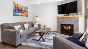 A living room with a grey sofa, a coffee table, a fireplace, and a flat-screen TV mounted above the fireplace. - Luxury 2Br/2Ba Townhouse- Pool, HT, Secure bike storage (Whistler)
