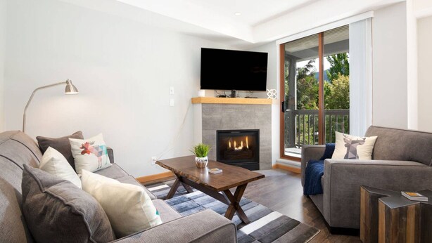 A modern living room with a fireplace, a flat-screen TV mounted above it, and a large window providing a view of the outdoors. - Luxury 2Br/2Ba Townhouse- Pool, HT, Secure bike storage (Whistler)