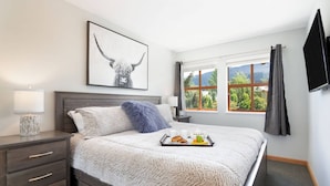 A bedroom with a large bed, a bedside table with a lamp, a framed picture on the wall, and a window with curtains. - Luxury 2Br/2Ba Townhouse- Pool, HT, Secure bike storage (Whistler)