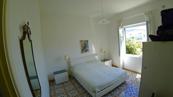 A bedroom with a bed, a nightstand, a lamp, a window with a view, and a wardrobe. - Mary's Romantic Terrace (Ischia)