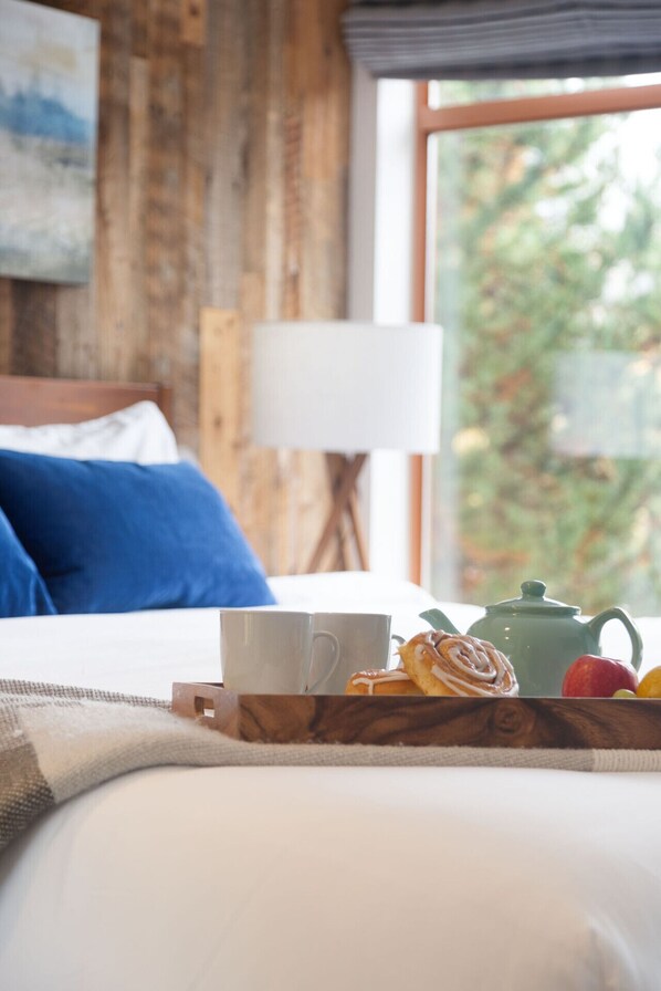 A neatly made bed with a tray of breakfast items including a teapot, a cup, and pastries, set against a wooden wall and a window with a view of trees.