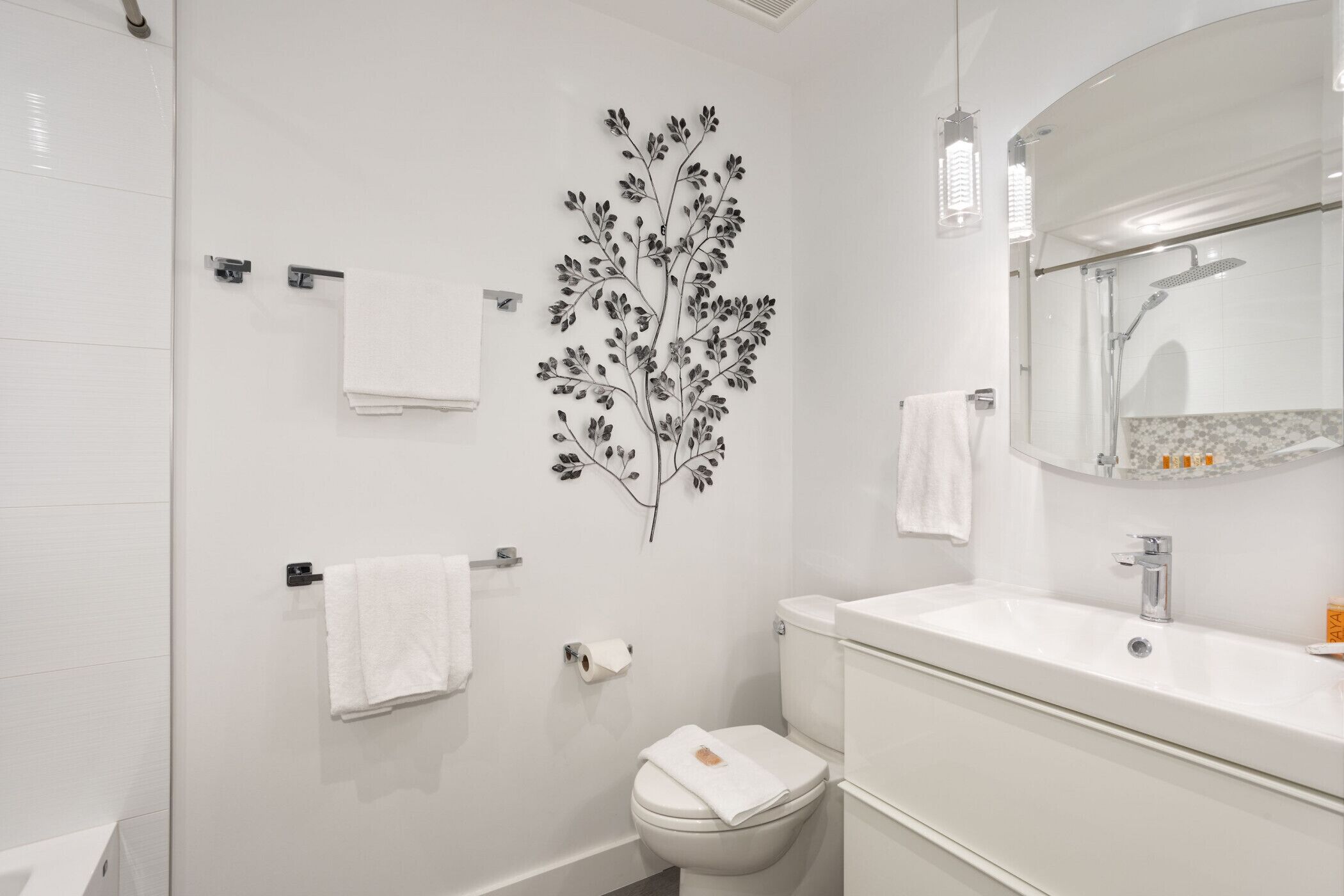 A modern bathroom with a white sink, toilet, and shower. There is a decorative wall art of a tree branch and a mirror above the sink.