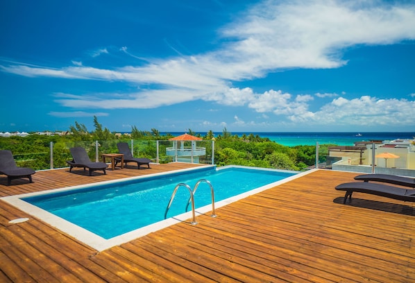 A wooden deck with a pool, lounge chairs, and a view of the ocean.