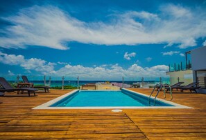 A wooden deck with a pool and lounge chairs, overlooking a beach and ocean.