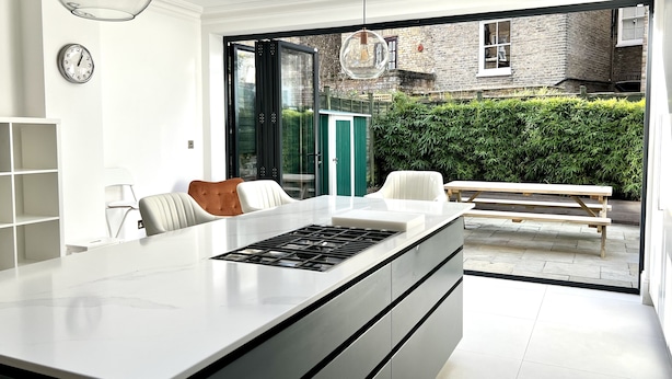 A modern kitchen with a large island, a dining area with a bench, and a view of a brick building and greenery outside. - Beautiful 4 bedroom Victorian family house on quiet road (London)