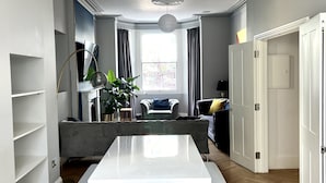 A modern dining area with a white table and chairs, a wooden floor, and a view of a living room with a sofa and a window. - Beautiful 4 bedroom Victorian family house on quiet road (London)