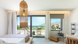 Une chambre d’hôtel moderne avec un grand lit, un banc en bois, une lampe en osier, une petite table et une vue sur la mer et les bâtiments.