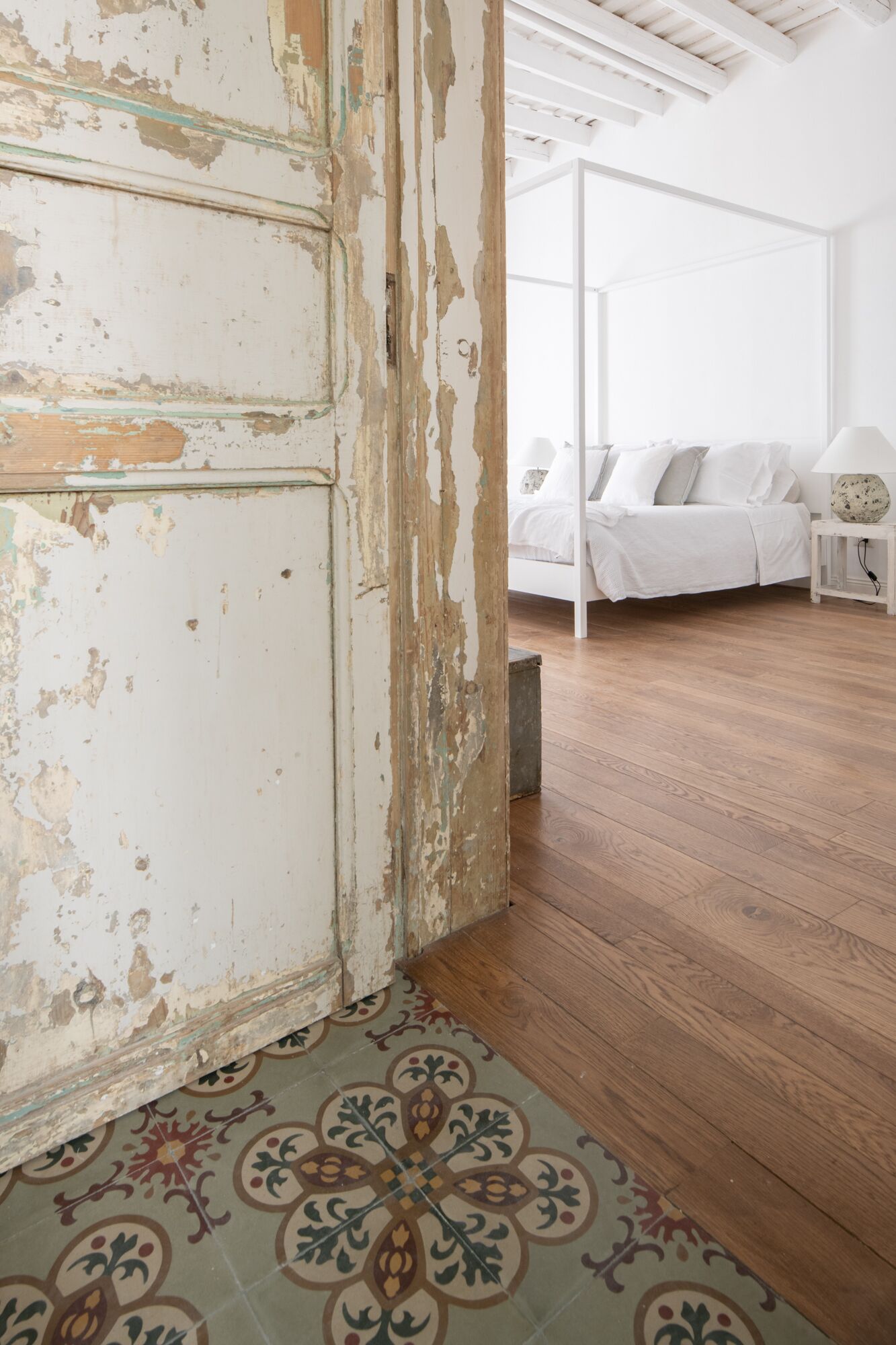 A room with a rustic, peeling white wall, a patterned floor, a wooden floor, a white bed with pillows, a bedside table with a lamp, and a white ceiling with wooden beams.