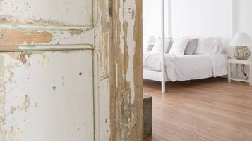 A room with a rustic, peeling white wall, a patterned floor, a wooden floor, a white bed with pillows, a bedside table with a lamp, and a white ceiling with wooden beams.