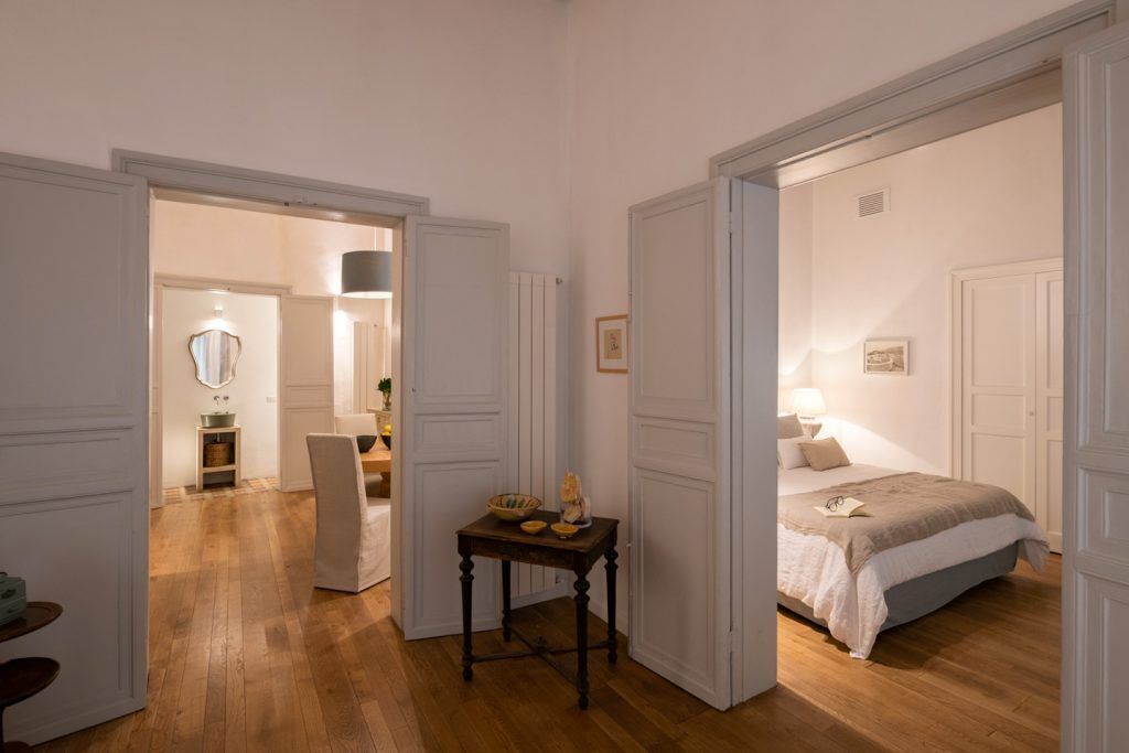 A hotel room with a wooden floor, a bed with a beige bedspread, a small table with a bowl and a bottle, and a chair.