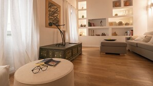 A living room with a sofa, ottoman, and a round table with glasses and books. There is a painting on the wall and a shelf with decorative items. - Dimora Botteghelle (Trapani)