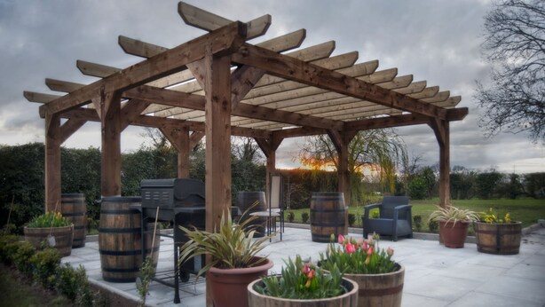 Une pergola en bois avec des sièges, des plantes en pot et un barbecue.