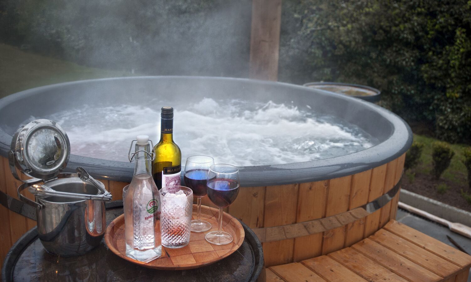 A hot tub with a wooden deck, a metal pot, a bottle, and two glasses filled with water.