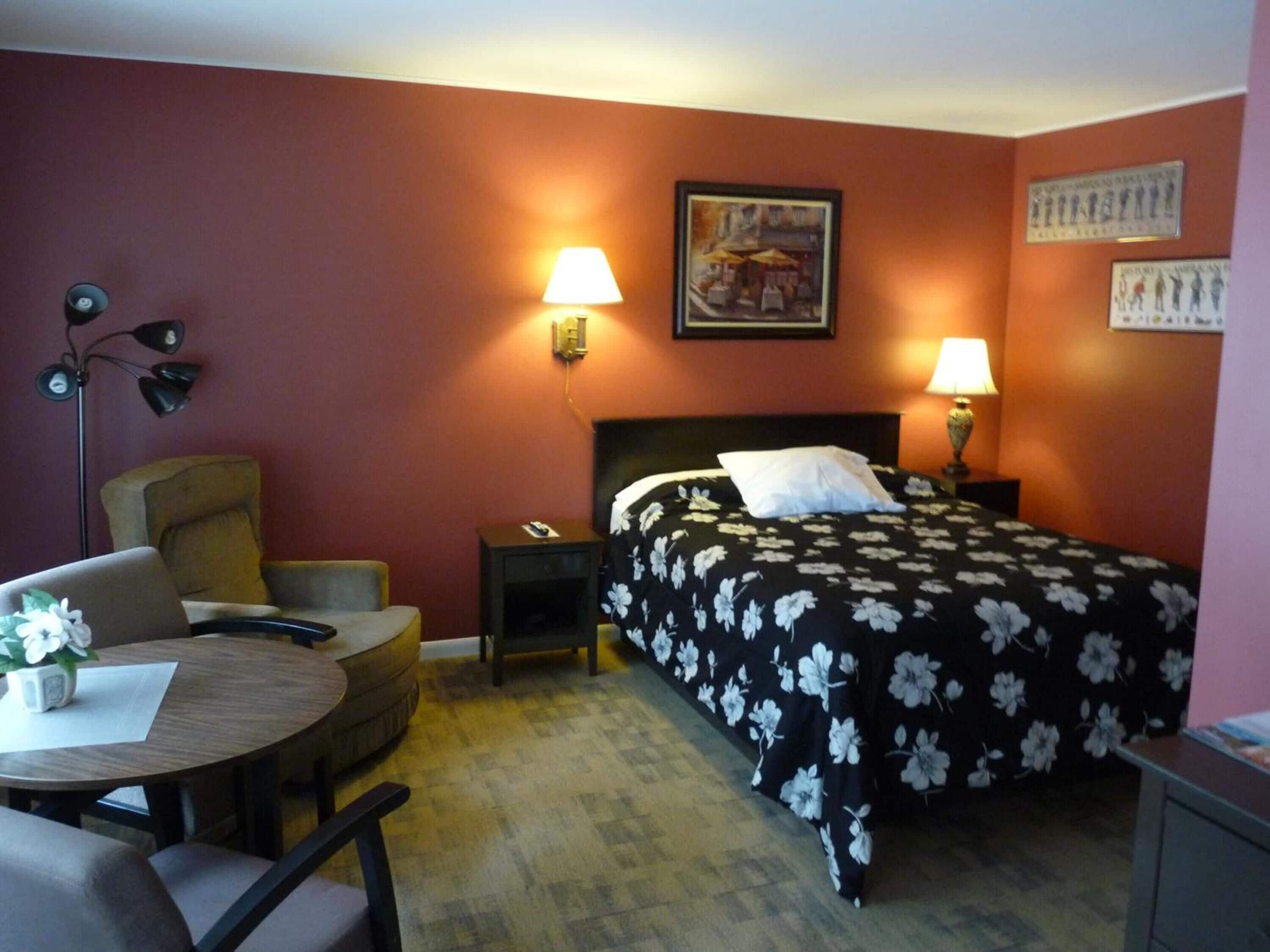 A hotel room with a bed, two bedside lamps, a desk with a chair, a small table with a vase, and a wall-mounted lamp.