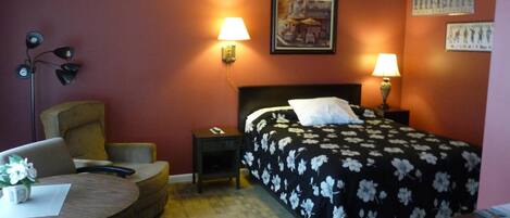 A hotel room with a bed, two bedside lamps, a desk with a chair, a small table with a vase, and a wall-mounted lamp.